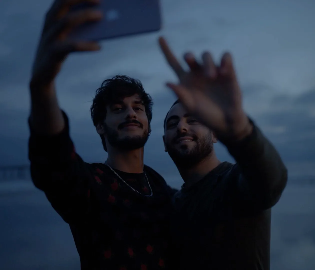 Two men taking a selfie together at dusk, one waving at the camera.
